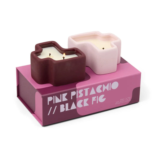 Two candles in decorative holders on a pink box labeled 'Pink Pistachio // Black Fig'.