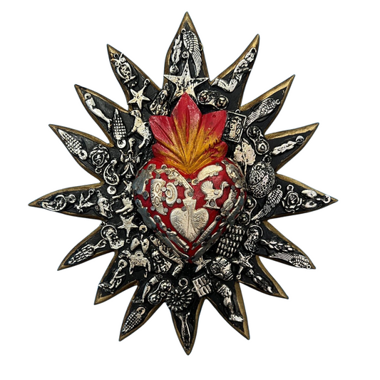 Wooden Mexican wall decor- Black Sun shape with a central red heart motif. The ornament is covered in charms which have a spiritual meaning. The decor piece is on a white background.
