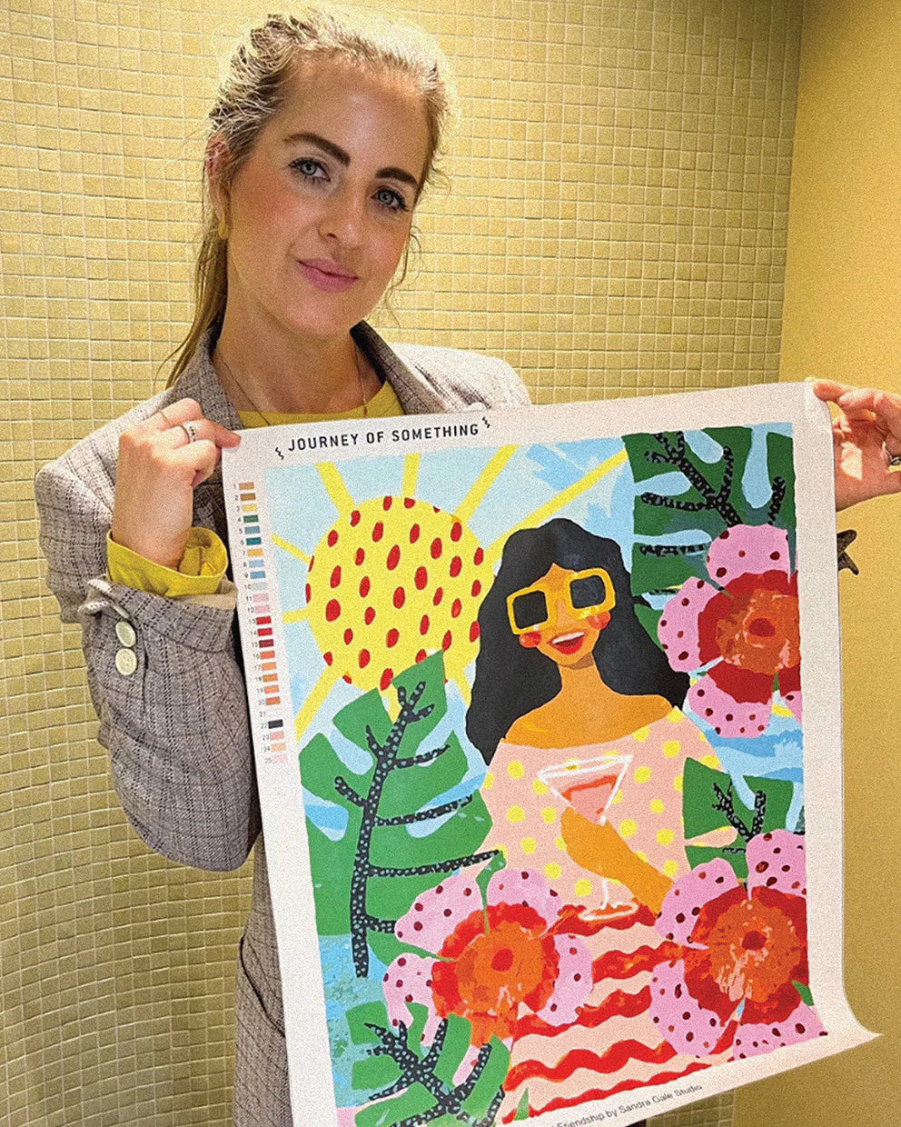 Person holding a colorful artwork with a woman, sun, and flowers on a yellow tiled wall background.