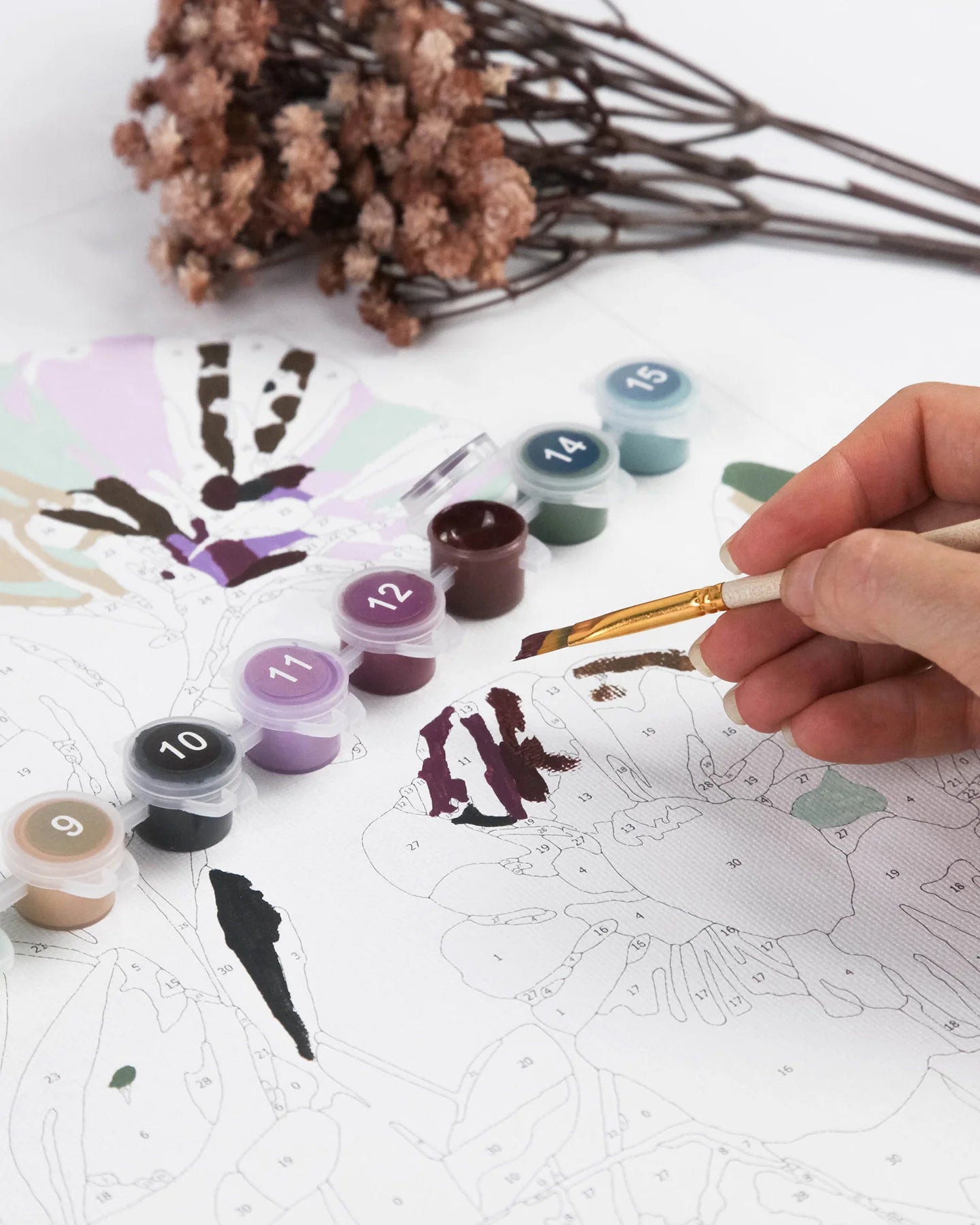 Person painting on a design with watercolor paints and a brush, surrounded by dried flowers.