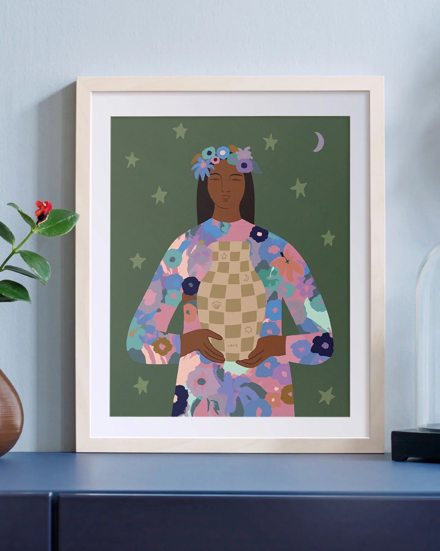 Finished paint by numbers of "Love Vase"Framed artwork of a woman with floral attire on a shelf against a light blue wall.