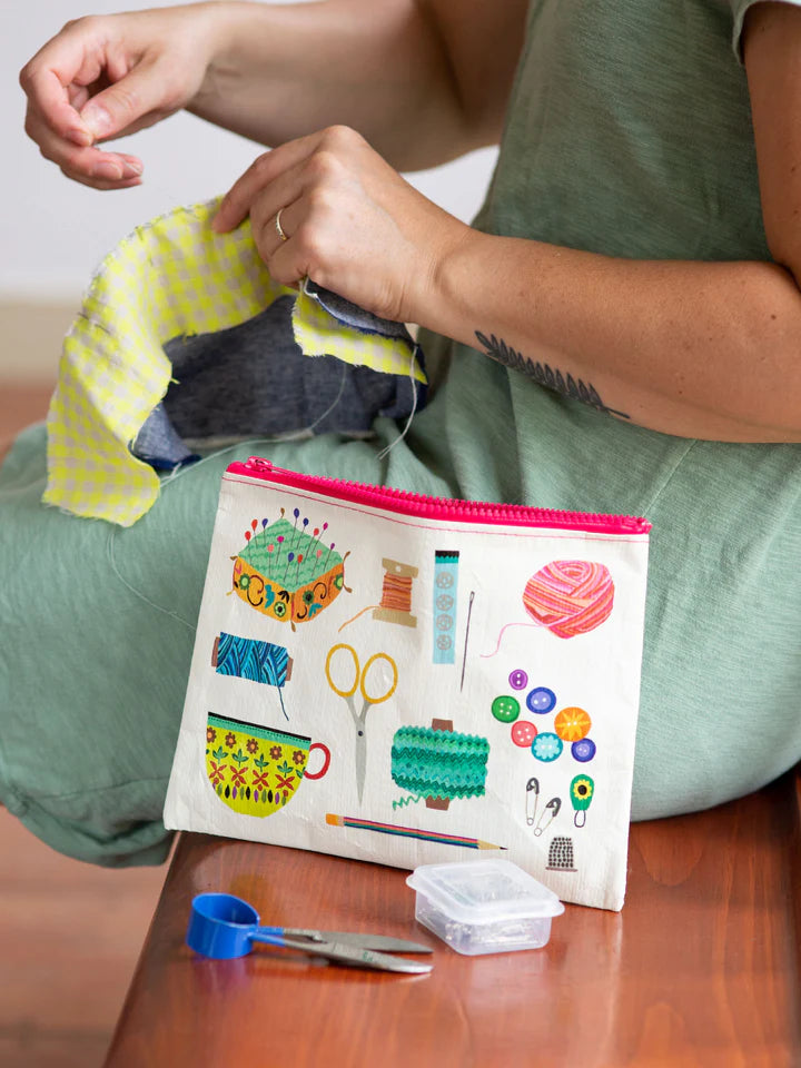 A woman seated  is hand sewing and her Blue Q  sewing kit print zipper pouch placed beside her. A pair of snippers and pin box are placed beside it.