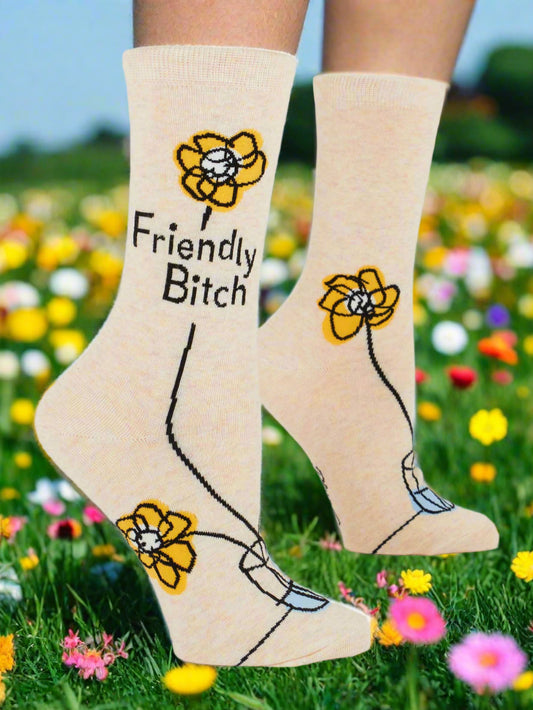 A creme pair of women's crew socks with a flower patter and "Friendly Bitch" slogan. The background is a field of flowers.