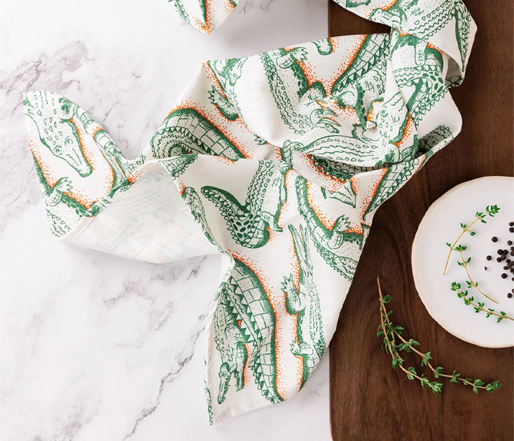 a scattered crocodile print tea towel on a marble table , a wooden side table featuring a plate and lemon myrtle twigs
