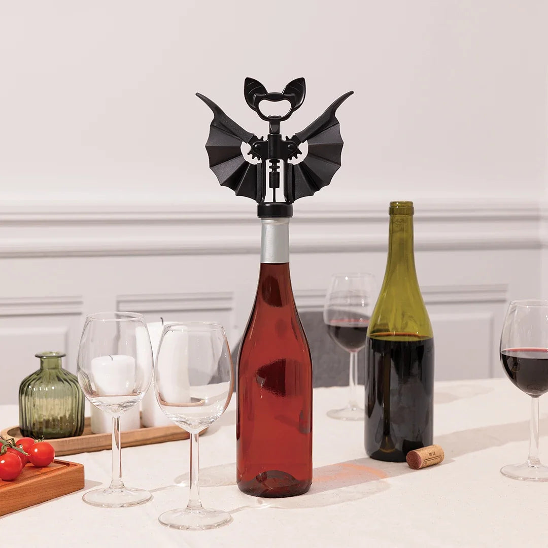 A bat shaped corkscrew on a bottle of red wine surrounded by glasses and another bottle of red wine on a white background setting