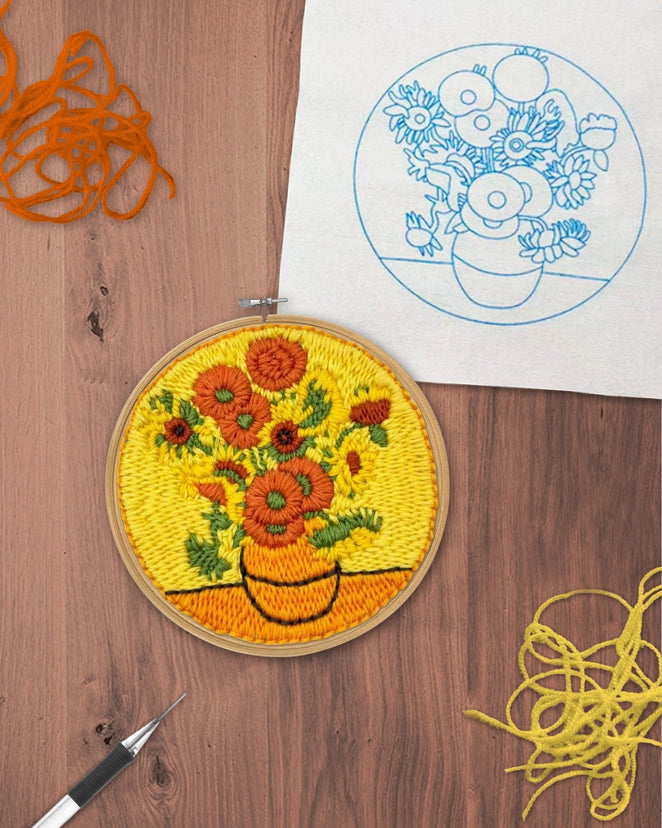 A completed embroidery hoop featuring a floral bouquet design in yellow, orange, and green thread, displayed on a wooden surface next to a blue line drawing of the same design on white fabric, with orange and yellow thread skeins nearby.