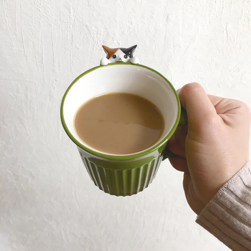 Hand holding a green mug with a cat-shaped handle against a white background