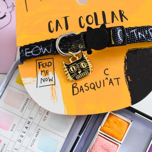 Cat collar with 'Feed Me Now' charm on a yellow card, placed on a surface with art supplies.