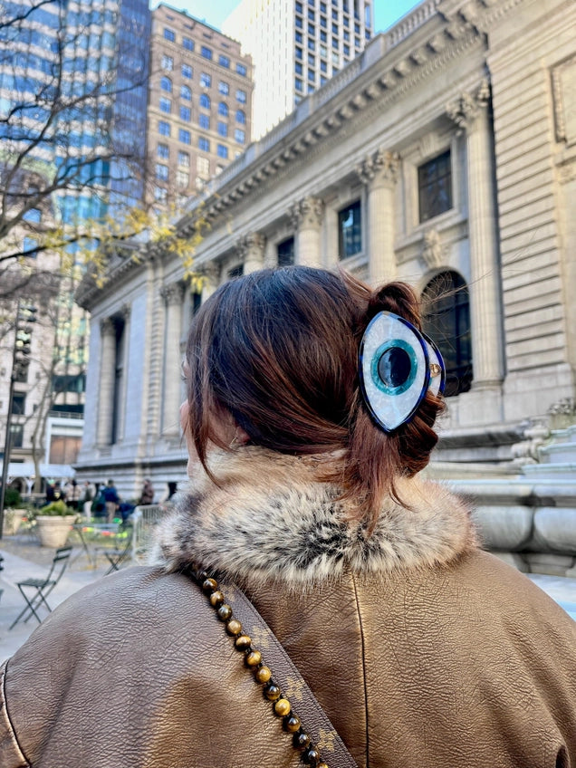 Person wearing a fur-lined coat with a decorative hair claw featuring an eye design, standing in an urban setting.