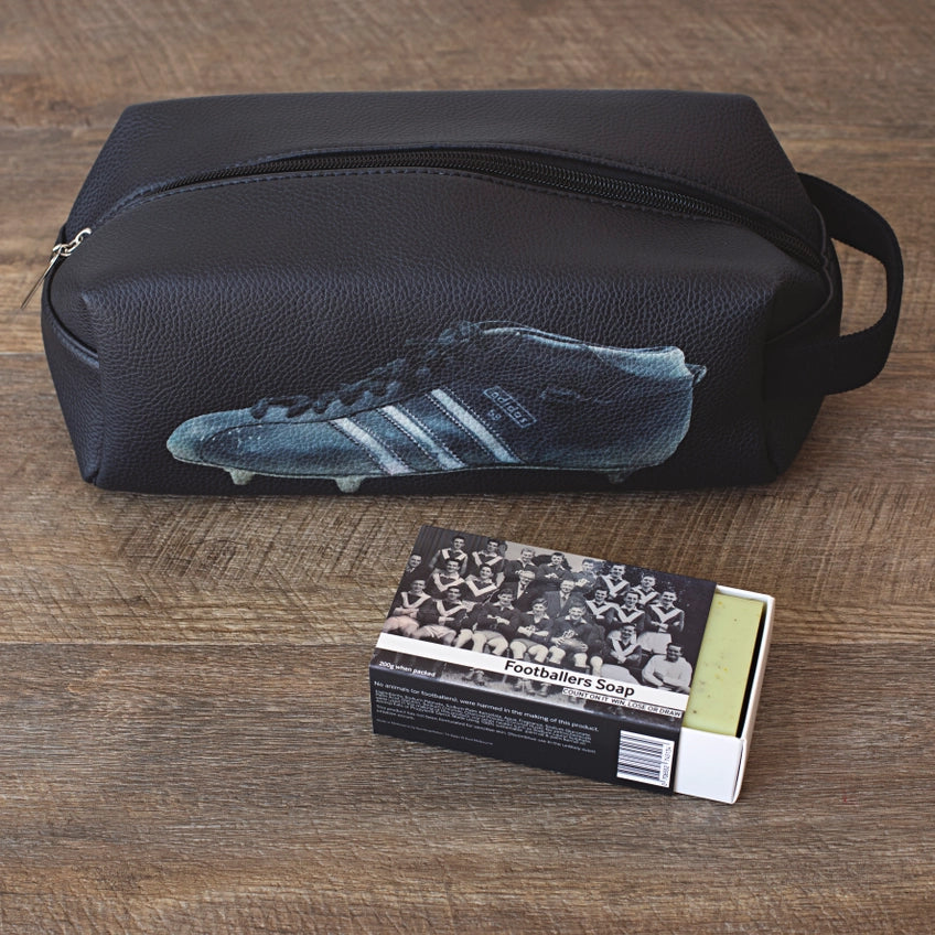 Black leather shoe bag with a football boot design on a wooden surface, next to a box of 'Footballers Soap'.