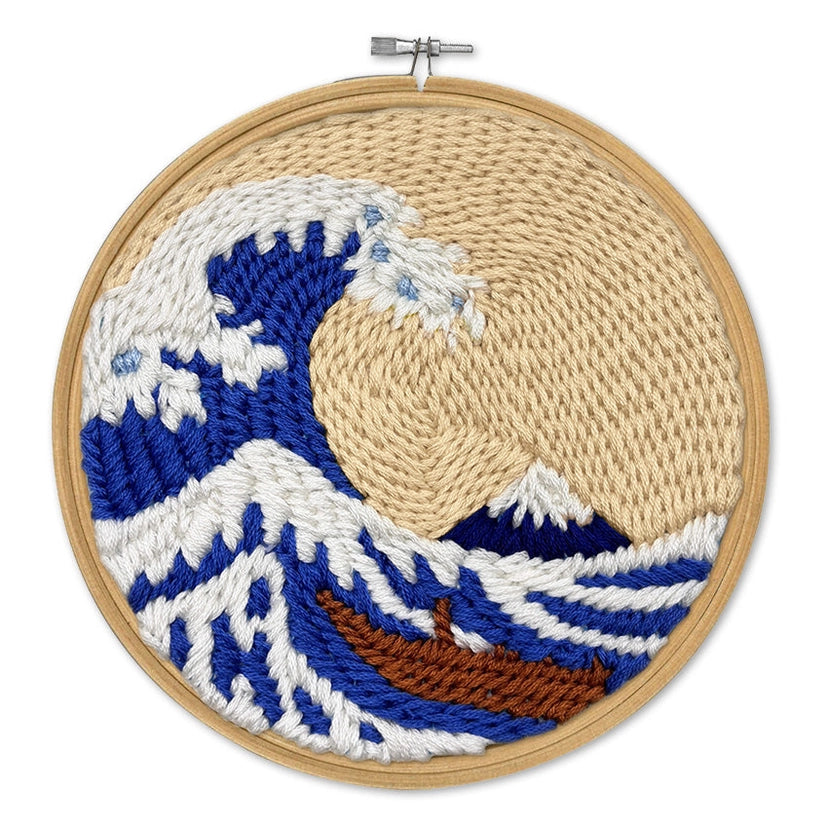 A circular wooden embroidery hoop containing a hand-embroidered design inspired by 'The Great Wave off Kanagawa'. The design features deep blue and white waves, a beige sun, a brown boat, and a white mountain, all set against a plain white background.