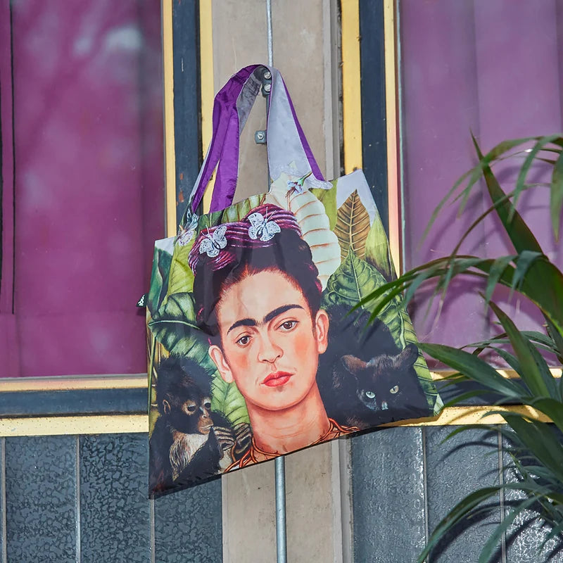 A shopping tote featuring Frida Kahlo hanging on a hook against a purple bacground.