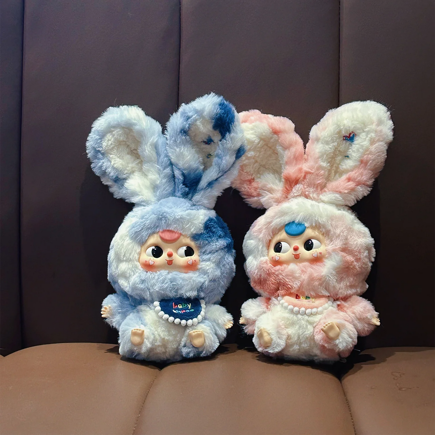 Two plush toys with bunny ears, one blue and one pink, sitting on a brown surface.