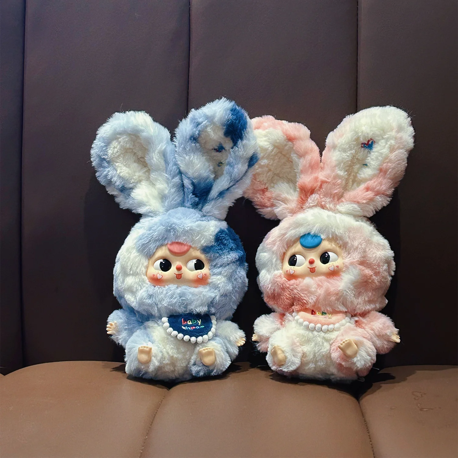 Two plush toys with bunny ears, one blue and one pink, sitting on a brown surface.