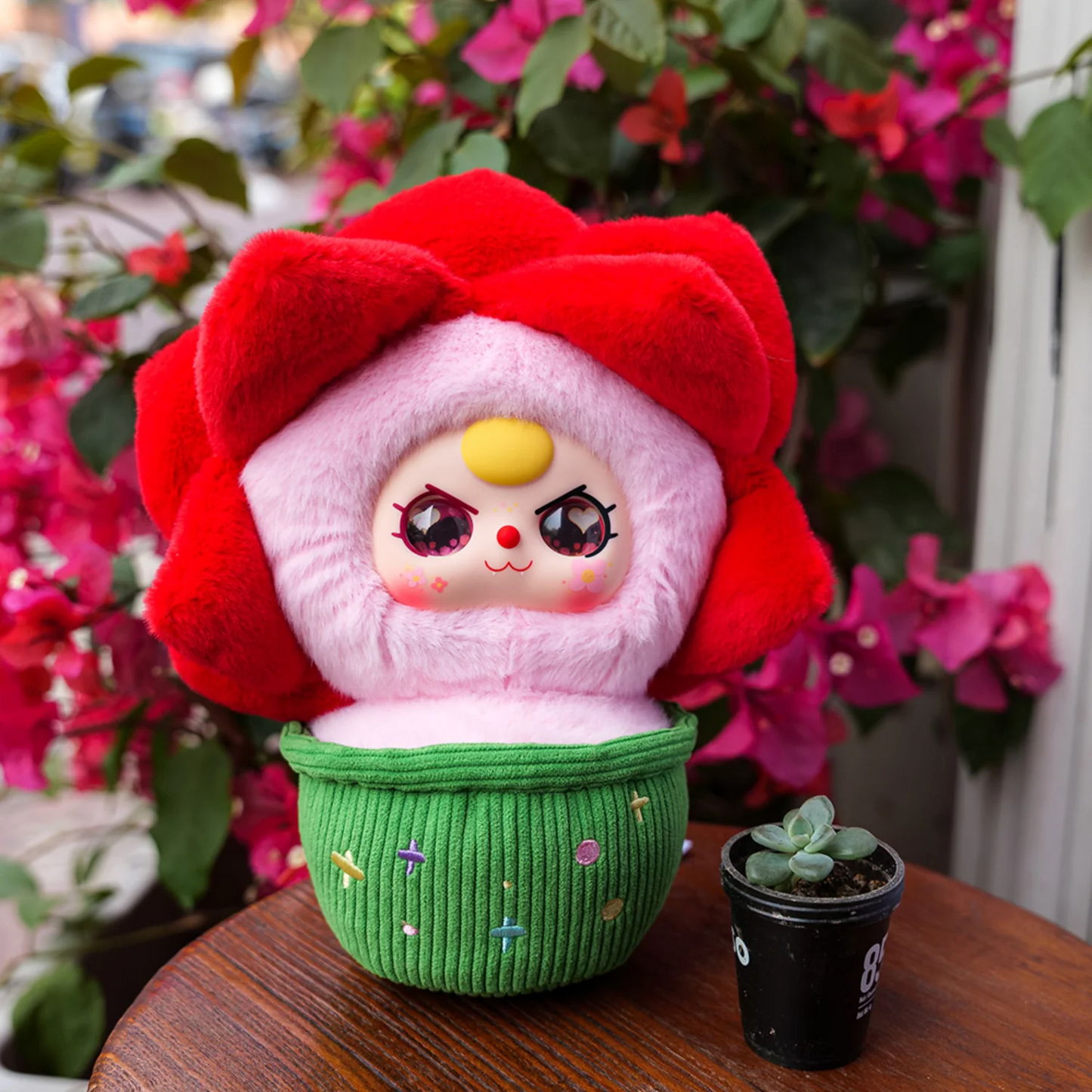 Plush toy with a red flower-like hat and pink face on a wooden surface with flowers in the background