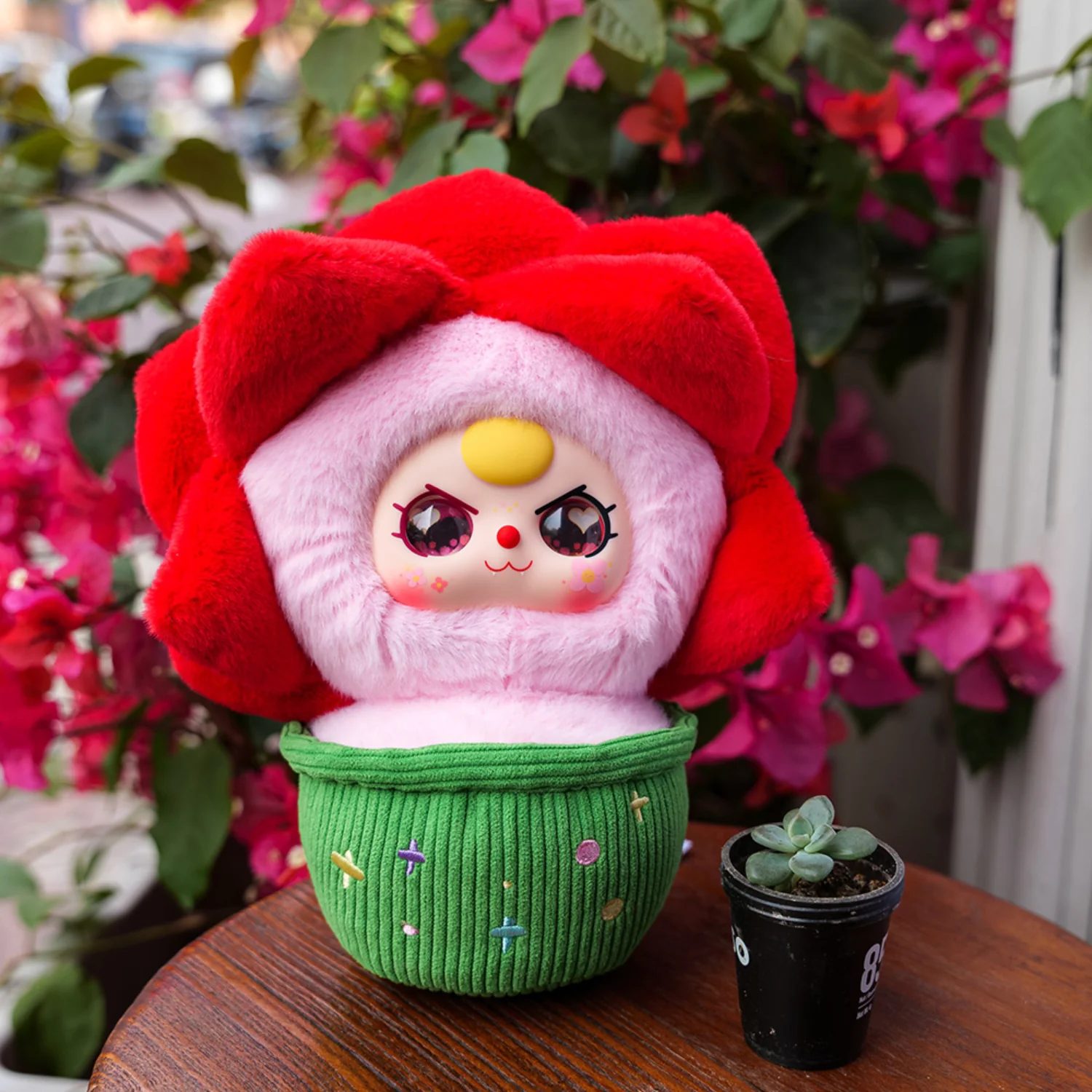 Plush toy with a red flower-like hat and pink face on a wooden surface with flowers in the background