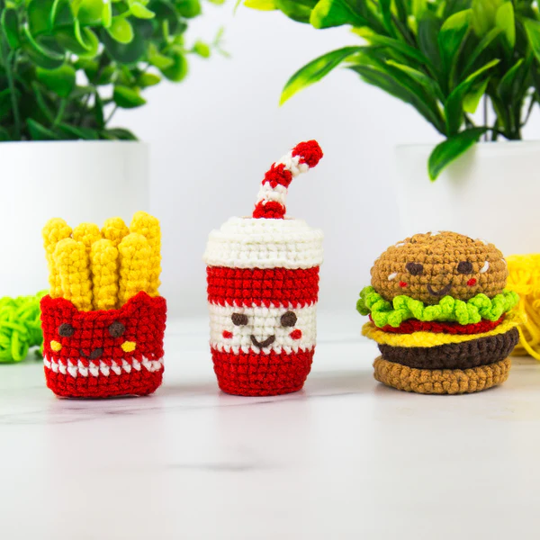 Three crocheted fast-food toys arranged on a white surface: a red and white french fries container with yellow fries, a red and white drink cup with a striped straw, and a brown burger with green and yellow details.