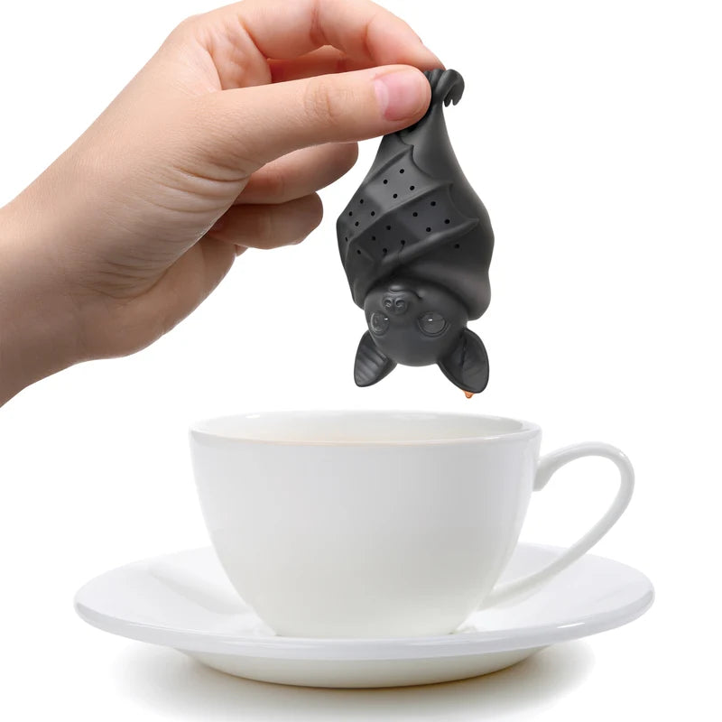 A hand holding a black bat tea infuser over a white teacup and saucer on a white backround.