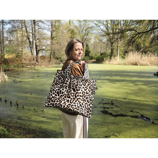 A woman overlooking a green wooded area and pond, looking back whilst holding  Leopard shopping bag over her shoulder.