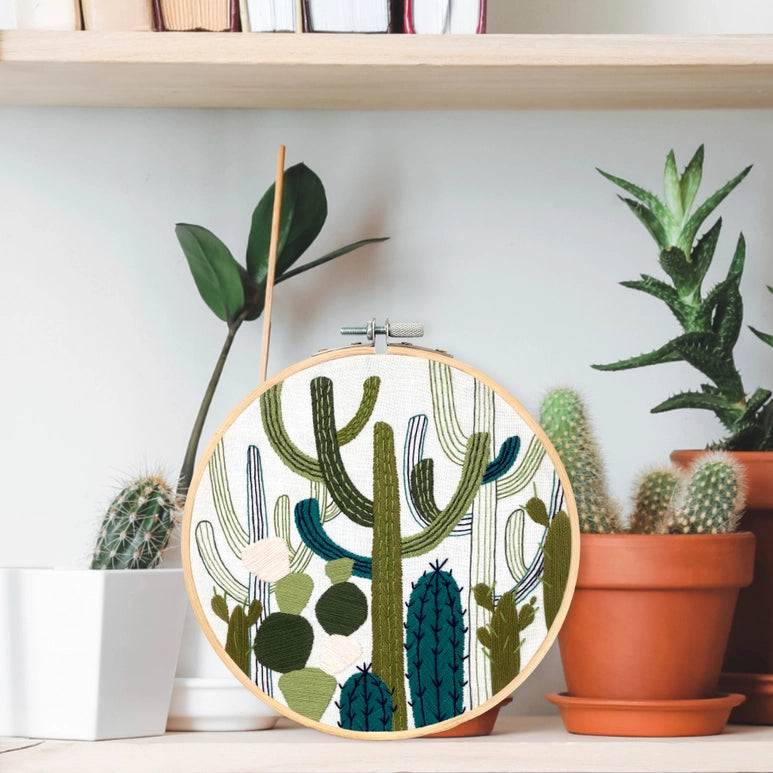 Lifestyle photo of a completed cactus garden embroidered 6" hoop. The loom is placed on a shelf amongst planters of cacti   