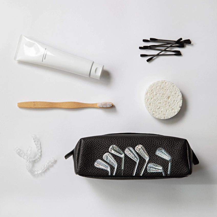 Black toiletry bag with white designs surrounded by a toothbrush, tube of toothpaste, cotton swabs, and sponge on a light gray background.