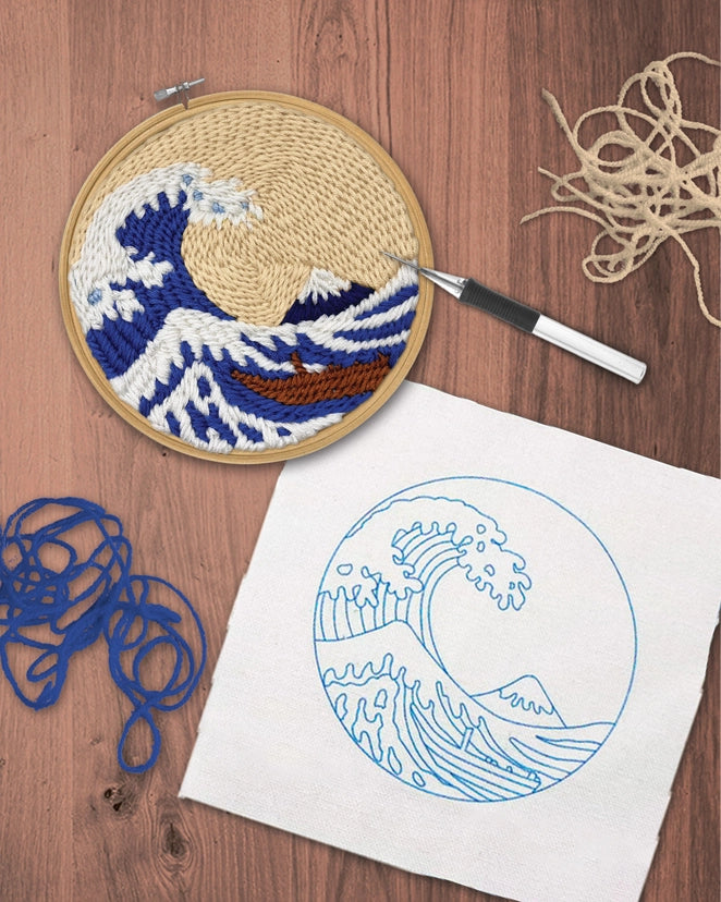 A circular embroidery hoop shows a wave-and-mountain design in beige, blue, white, and brown thread, placed on a wooden surface beside a white fabric sample with blue wave linework, a pen, and loose blue and beige thread.