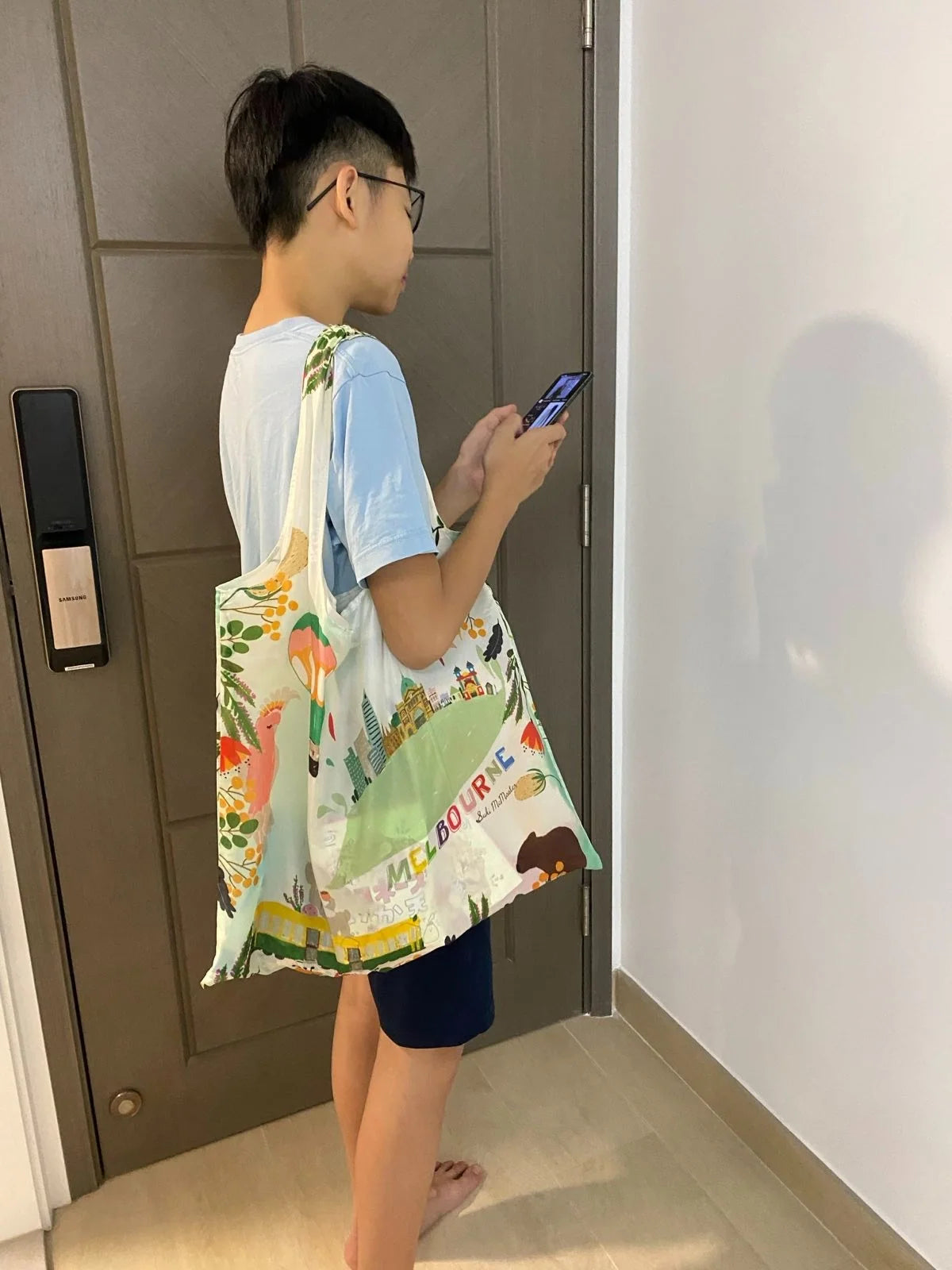 A young boy standing in front  khaki door, looking down at his phone and carrying a shopping bag with Melbourne icons
