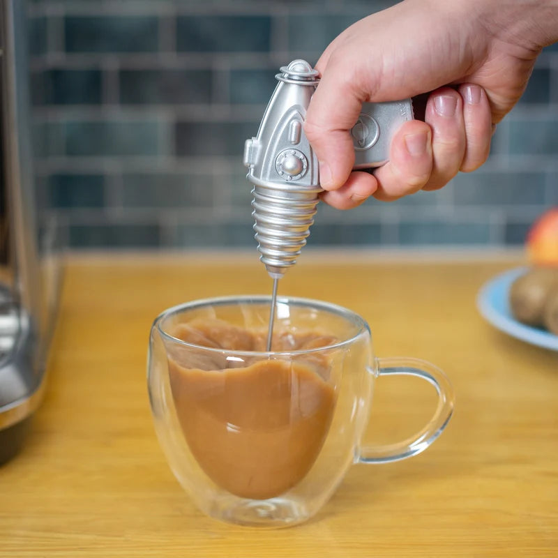 A hand using a space gun shaped milk frother in a cup of coffee placed on a wooden bench with a background of blue tiles