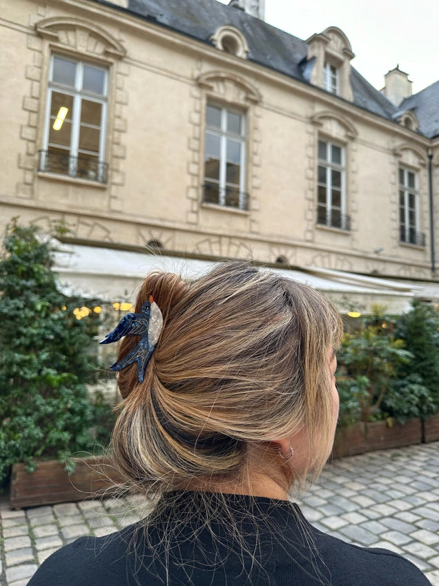 Person with styled hair in an outdoor setting with a building and plants in the background
