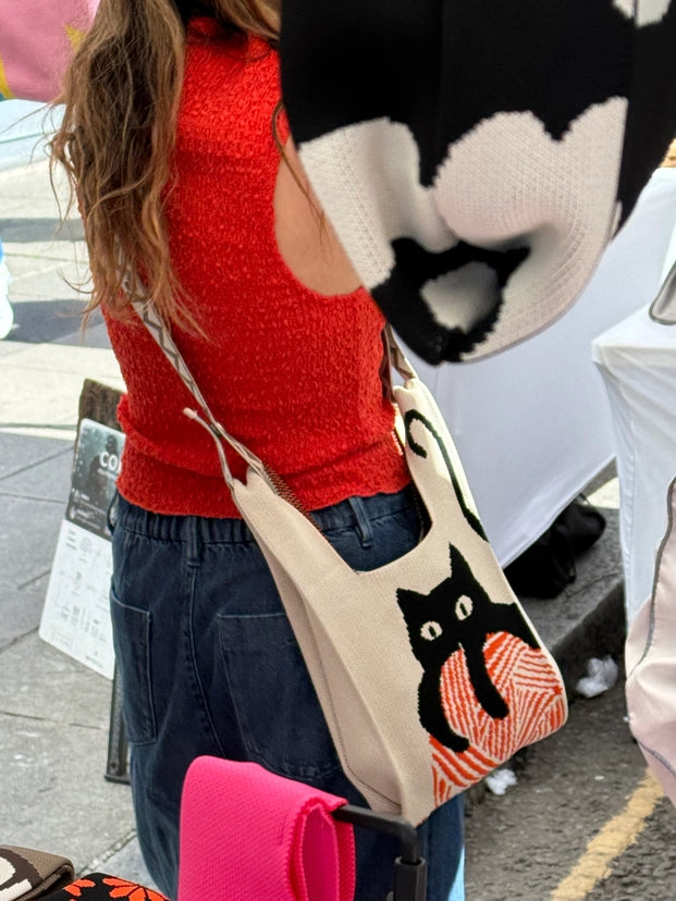 Person wearing a red sweater and black and white cow print hat, holding a beige bag with a black cat design.