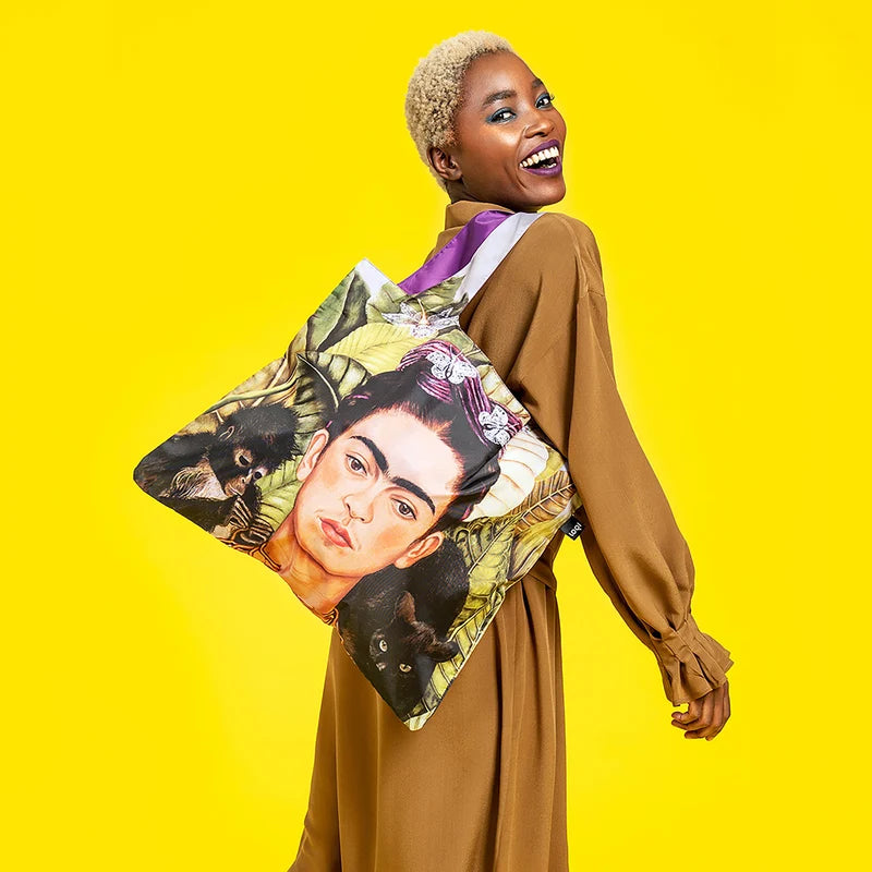 A smiling lady wearing a brown dress posing with a  Loqi Frida Kahlo shopping bag on a yellow background