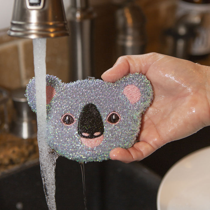 A hand holding a KIKKERLAND Koala sponge near a sink and running water